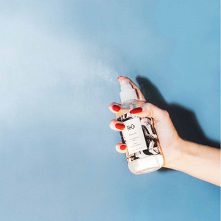 Hand holding a spray bottle labeled 'R+Co' against a blue background